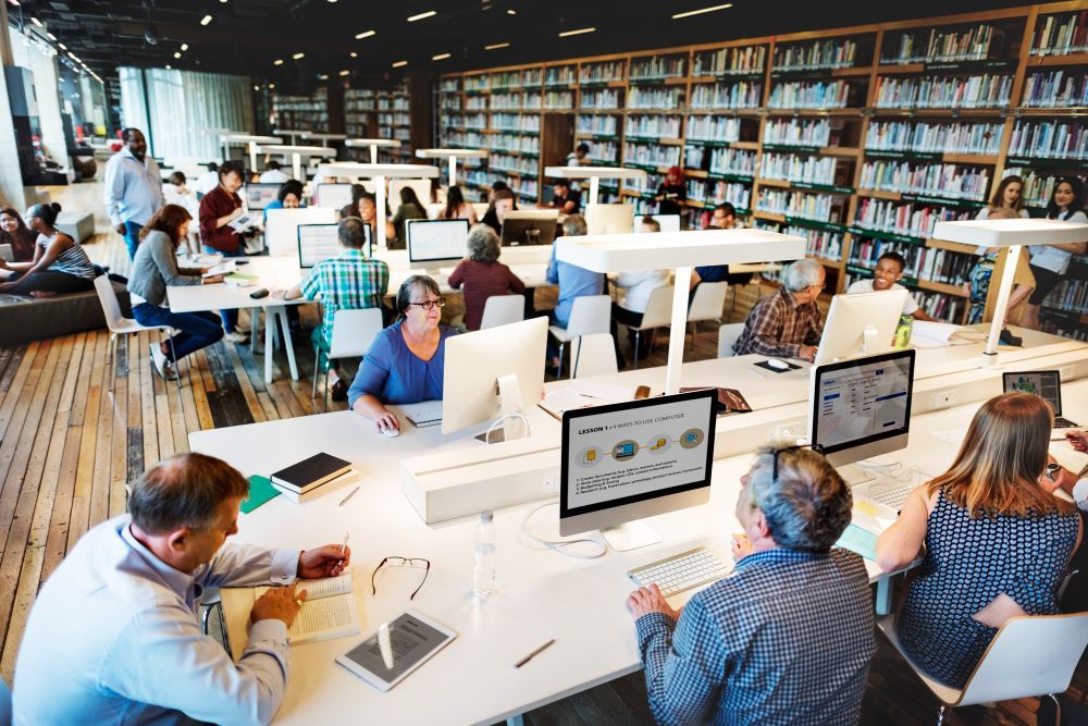 Busy library computer lab with people studying and working at desks amid bookshelves