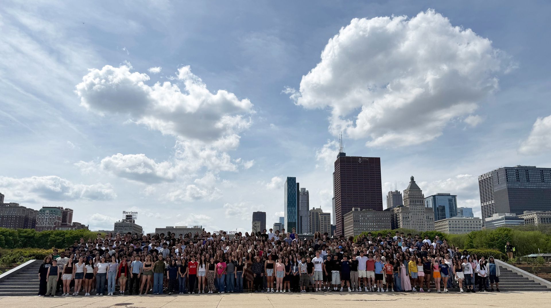Large crowd gathered on a city plaza with skyline buildings and cloudy sky in the background.