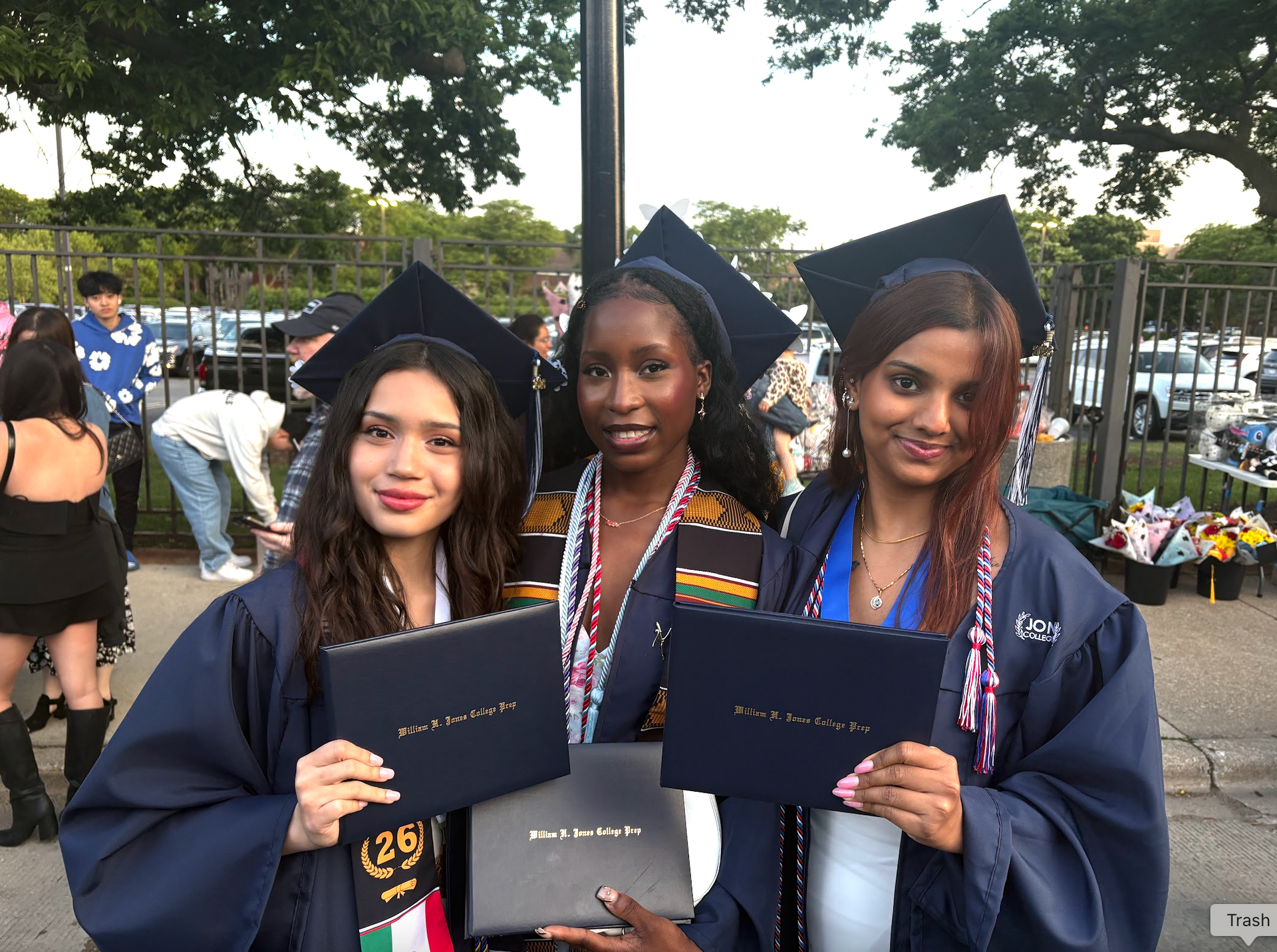 Three graduates in caps and gowns holding diplomas outdoors at a graduation ceremony