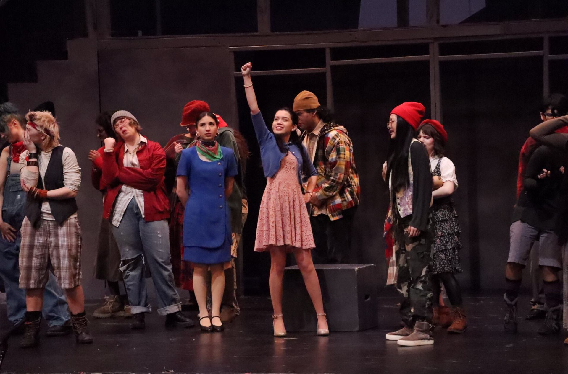 Costumed performers standing on a dark stage, one with an arm raised, others clustered in a line.