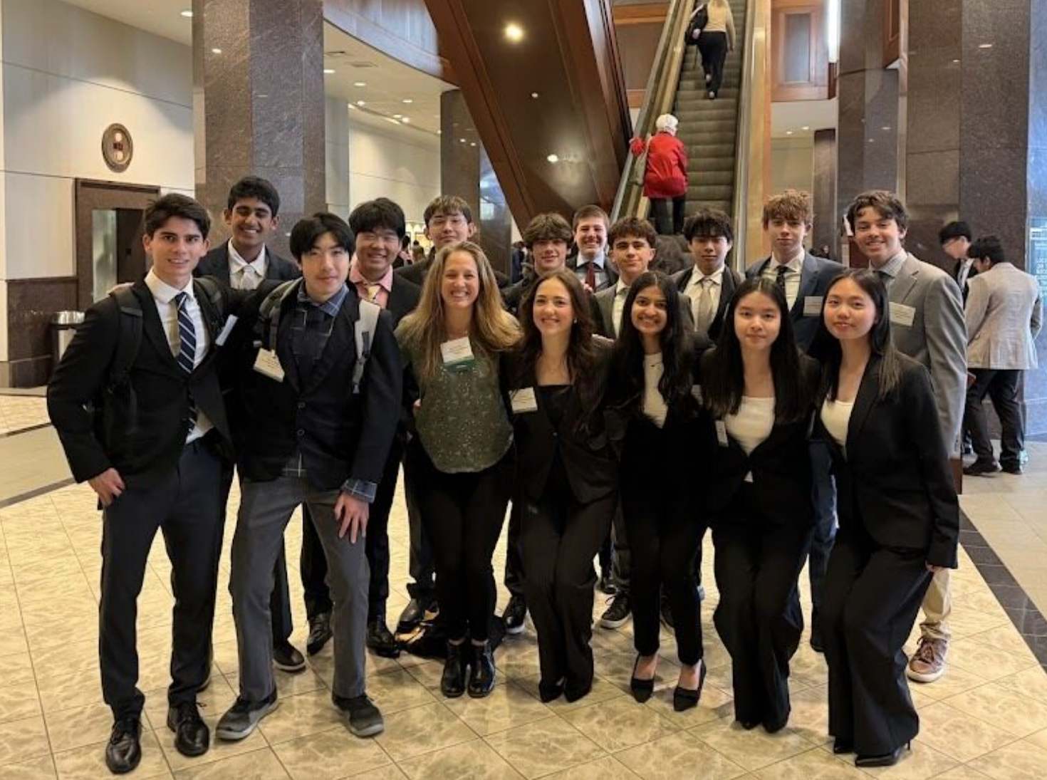 Group of people posing in formal attire in a bright indoor lobby with marble floors and escalators.