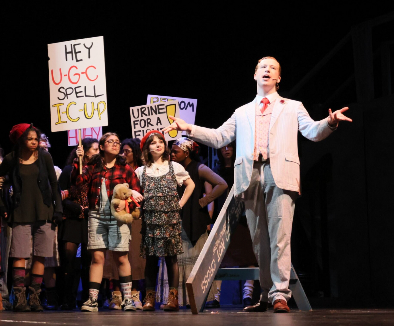 Stage protest scene with a man speaking beside students holding signs, including “HEY UGC SPELL ICU” and “UNION NOW”