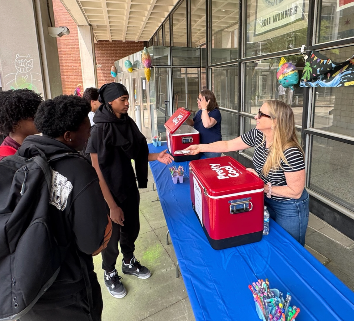 Students at a table with red coolers outside a storefront, serving drinks and snacks