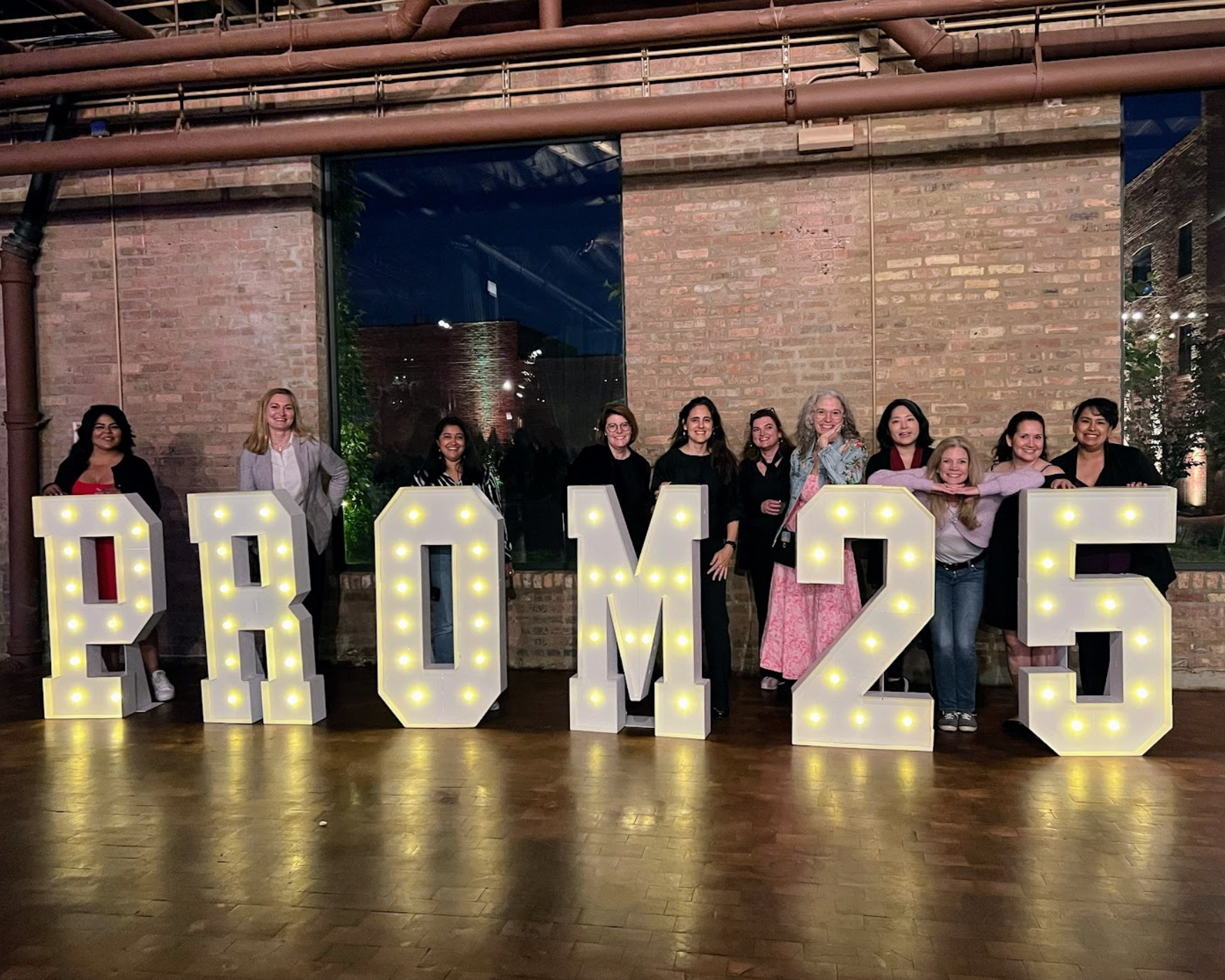 People posing behind lit marquee letters spelling “PROM25” in a rustic indoor venue.