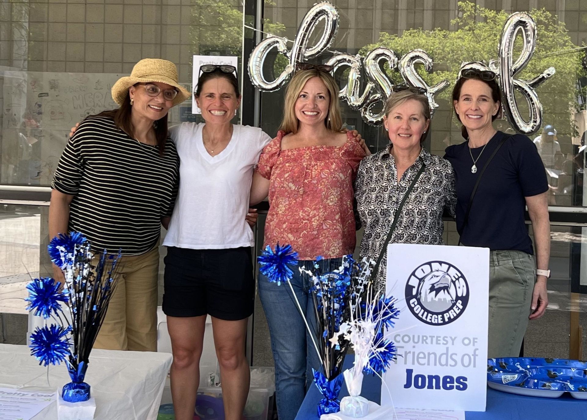 Five women posing at a Friends of Jones event with a sign and blue-and-white pom-poms