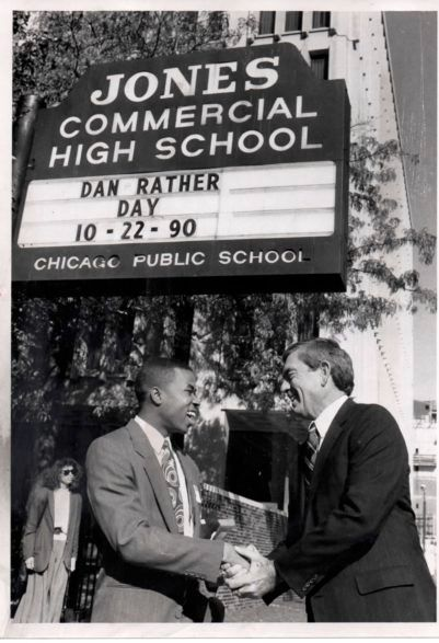 Two men shake hands beneath a Jones Commercial High School sign with a day and date board.