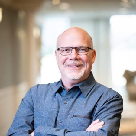 Man with glasses smiles, arms crossed, wearing a blue button-down shirt in a well-lit office.