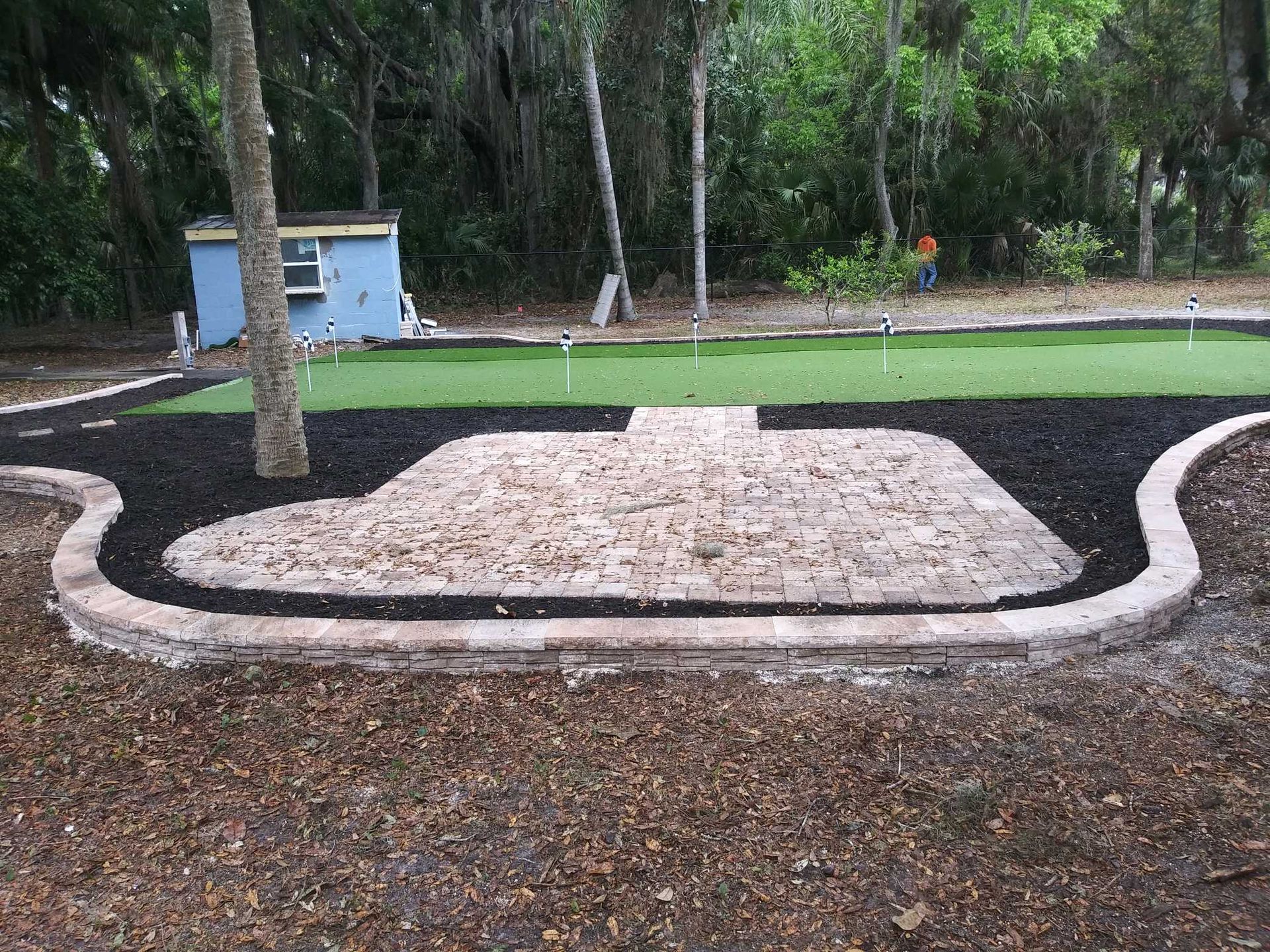 A paver patio with a curved stone border set against a backyard putting green with black mulch and a shed in the distance.