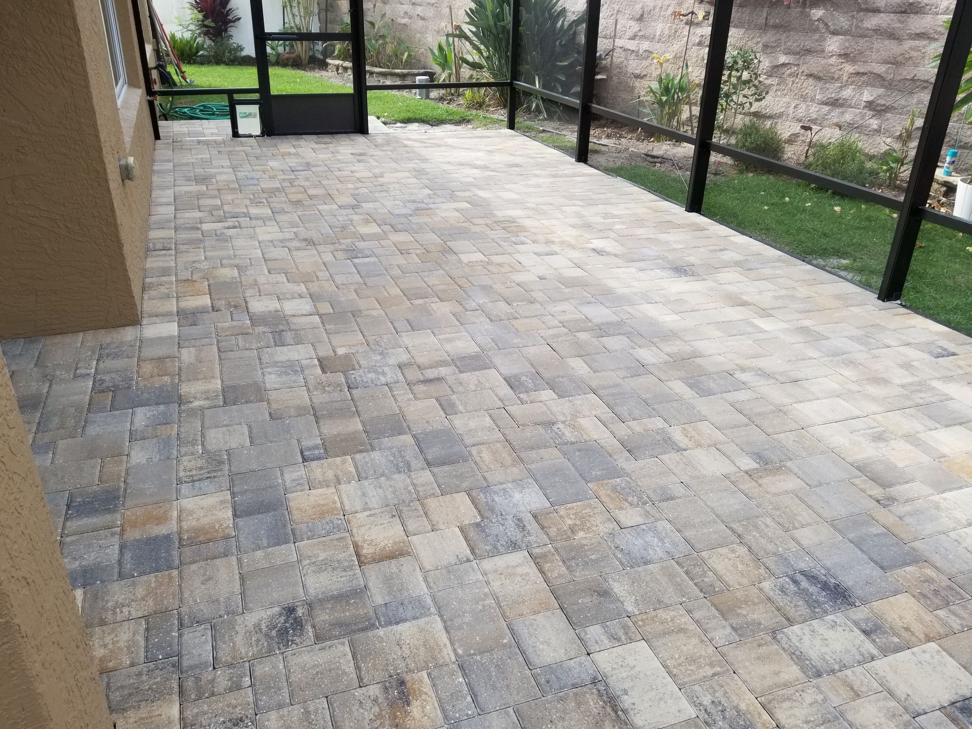 A patio made of multicolored rectangular stone pavers enclosed by a dark screened-in frame looking out toward a lawn.