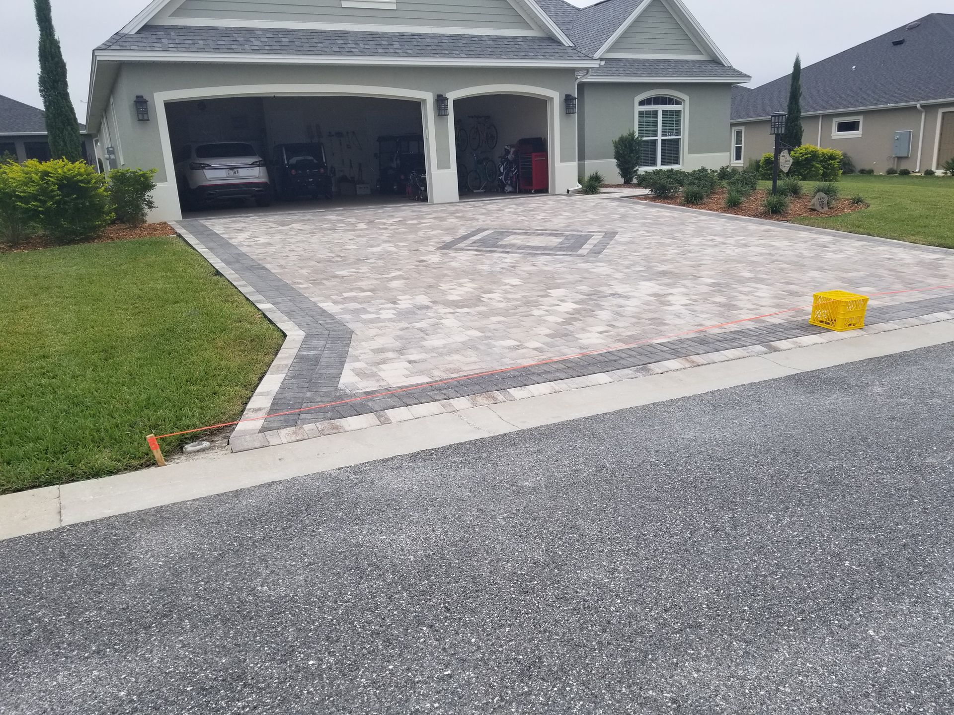 A residential driveway made of light patterned pavers with a dark border, leading to a two-car garage.