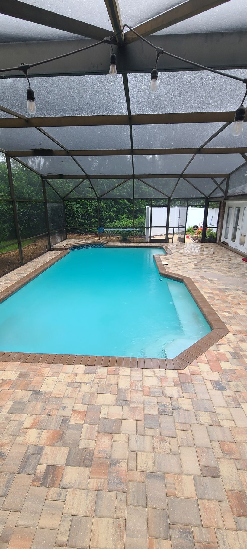 A rectangular turquoise swimming pool on a stone patio, enclosed by a screened-in lanai with hanging string lights.