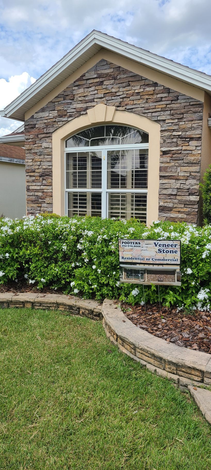 A tan home exterior features a stone-faced gable with an arched window, surrounded by white flowering bushes and lawn.