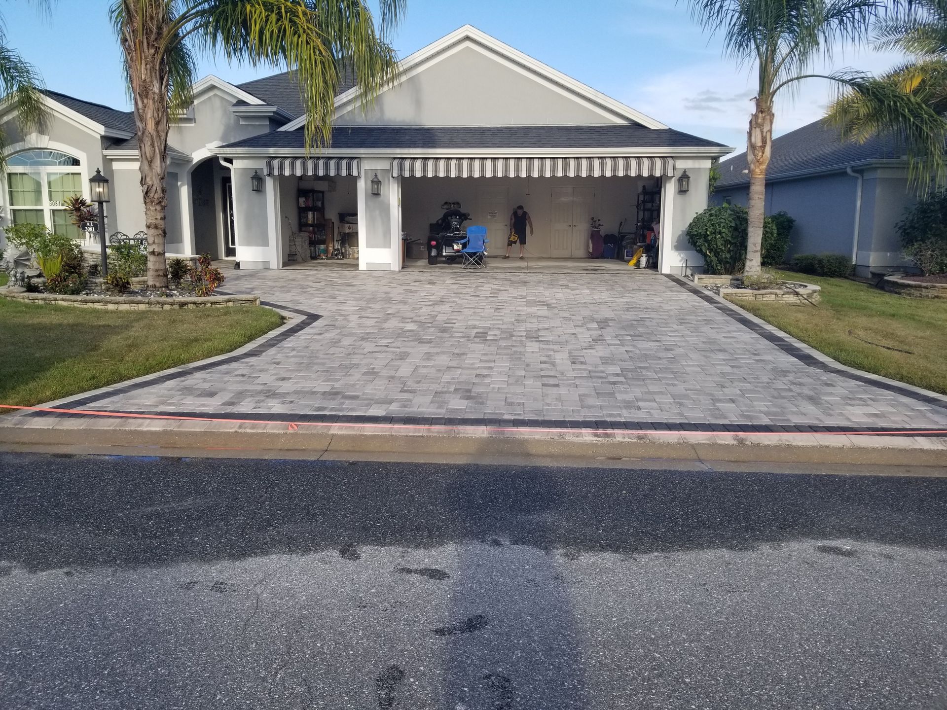 A house with a large, light-colored paver driveway leading to a three-car garage under a striped awning, on a sunny day.