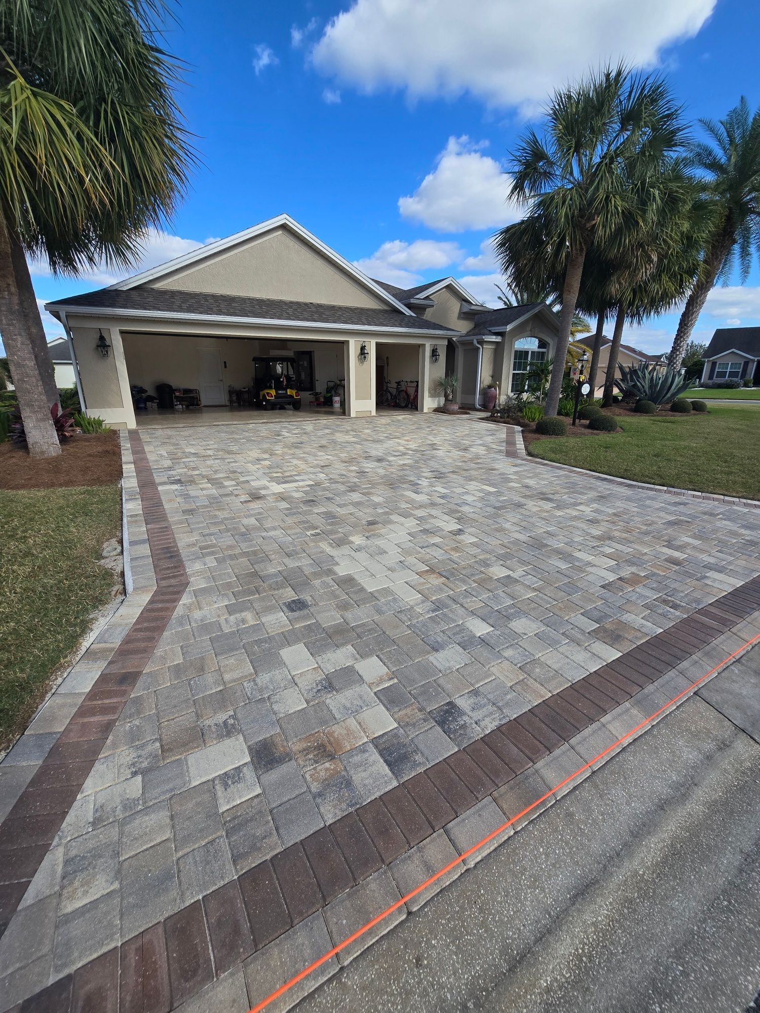 A suburban house with a newly installed patterned paver driveway featuring a dark red border, set under a blue sky.