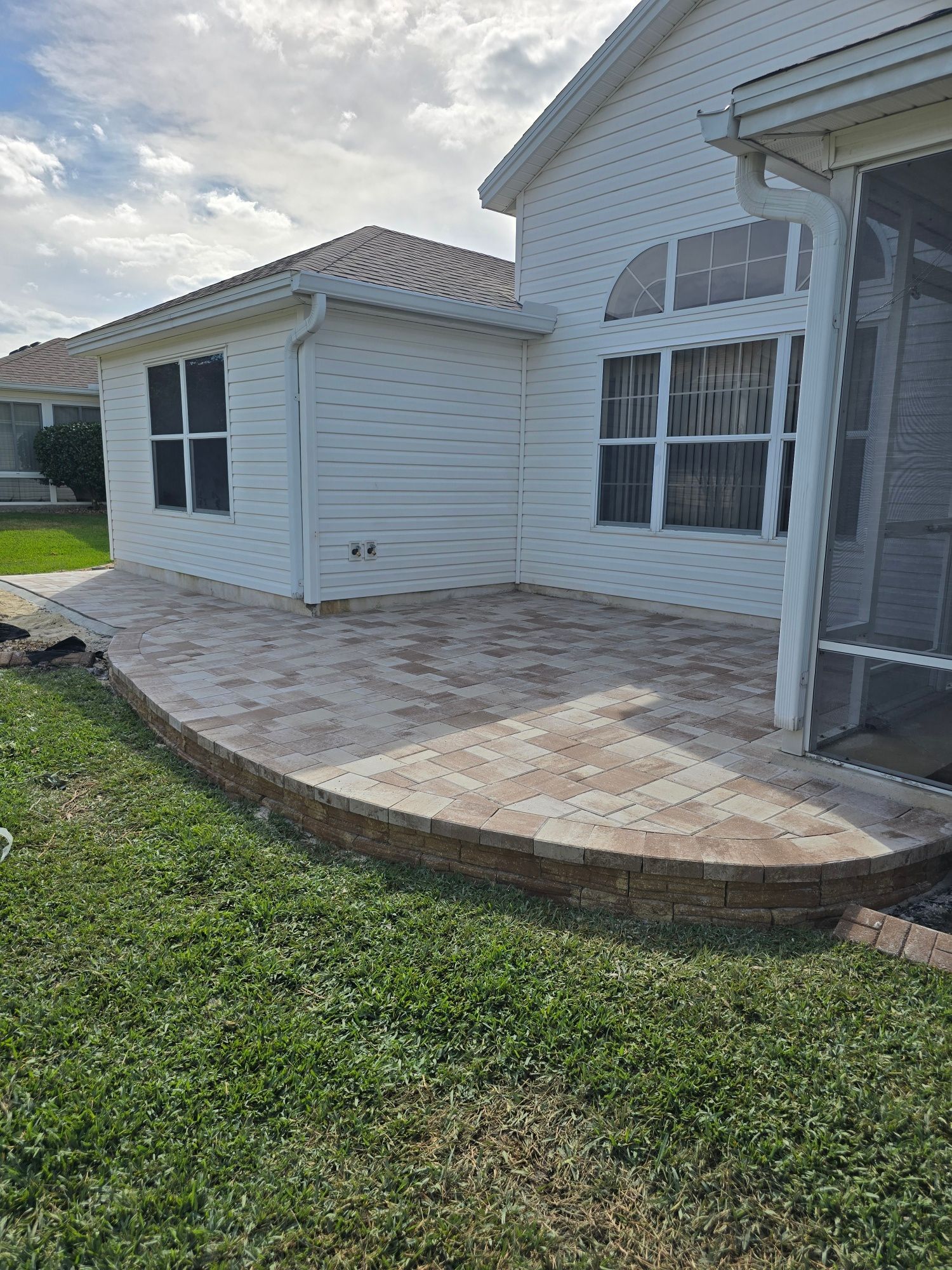 A newly installed paver patio with a curved retaining wall extending from the side of a white suburban house.