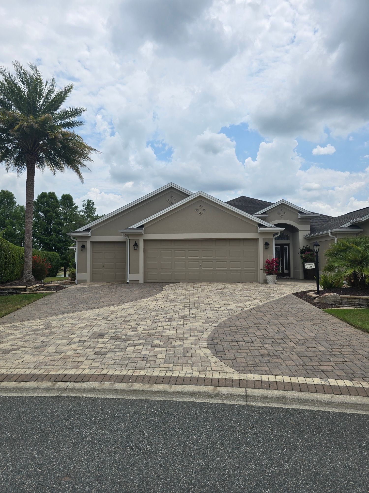 A beige single-story house with a decorative paver driveway and a tall palm tree under a cloudy sky.