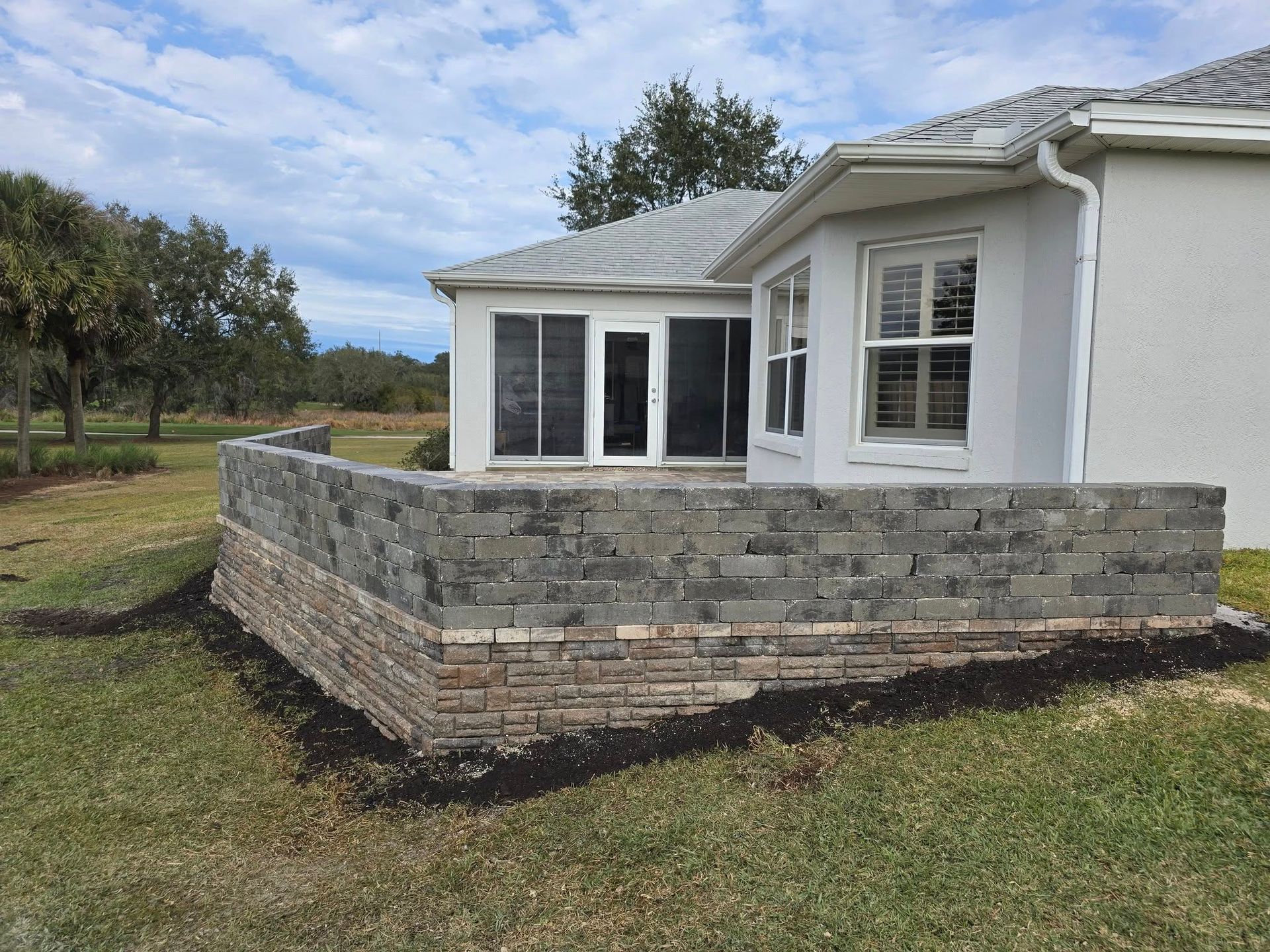 A stone retaining wall built in front of a white house with screened-in windows, set against a grassy landscape.