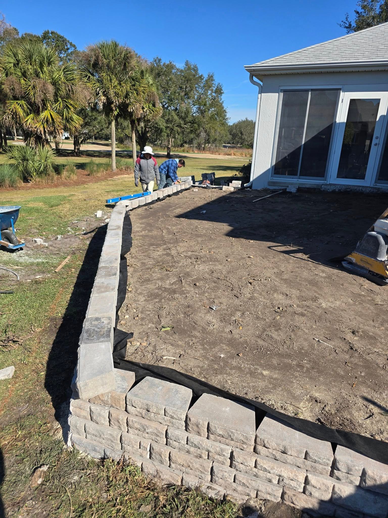Workers build a gray stone retaining wall for a patio alongside a white house on a sunny day.