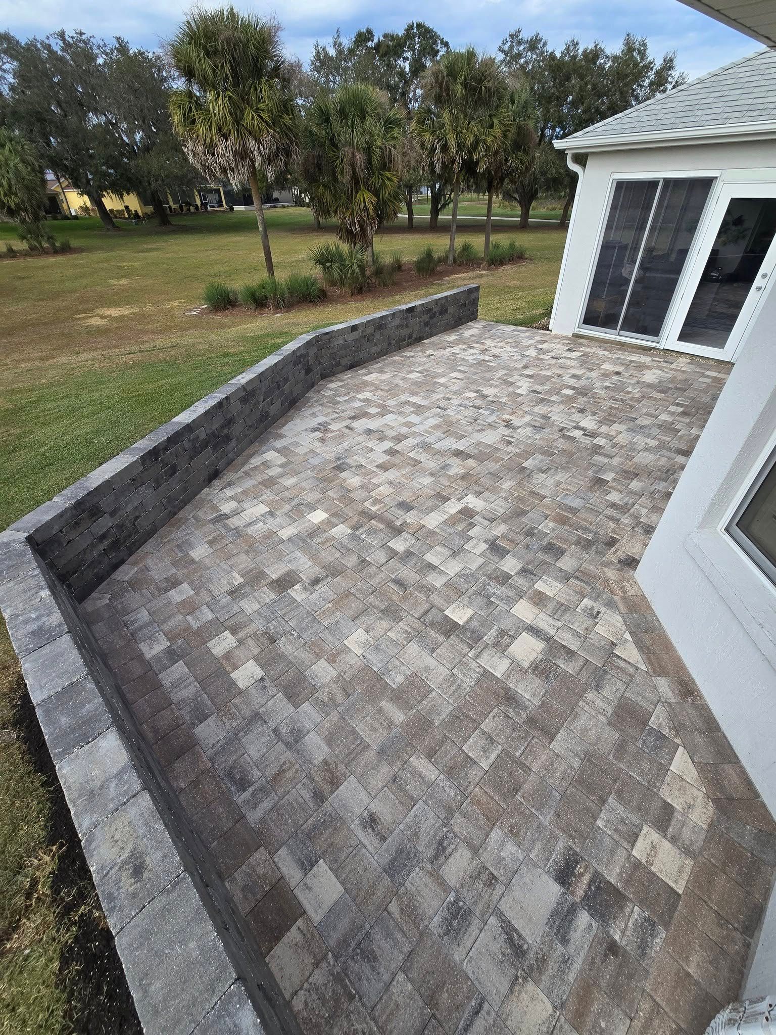 A light-colored stone paver patio featuring a multi-sided, dark gray retaining wall, bordered by a grassy lawn and trees.