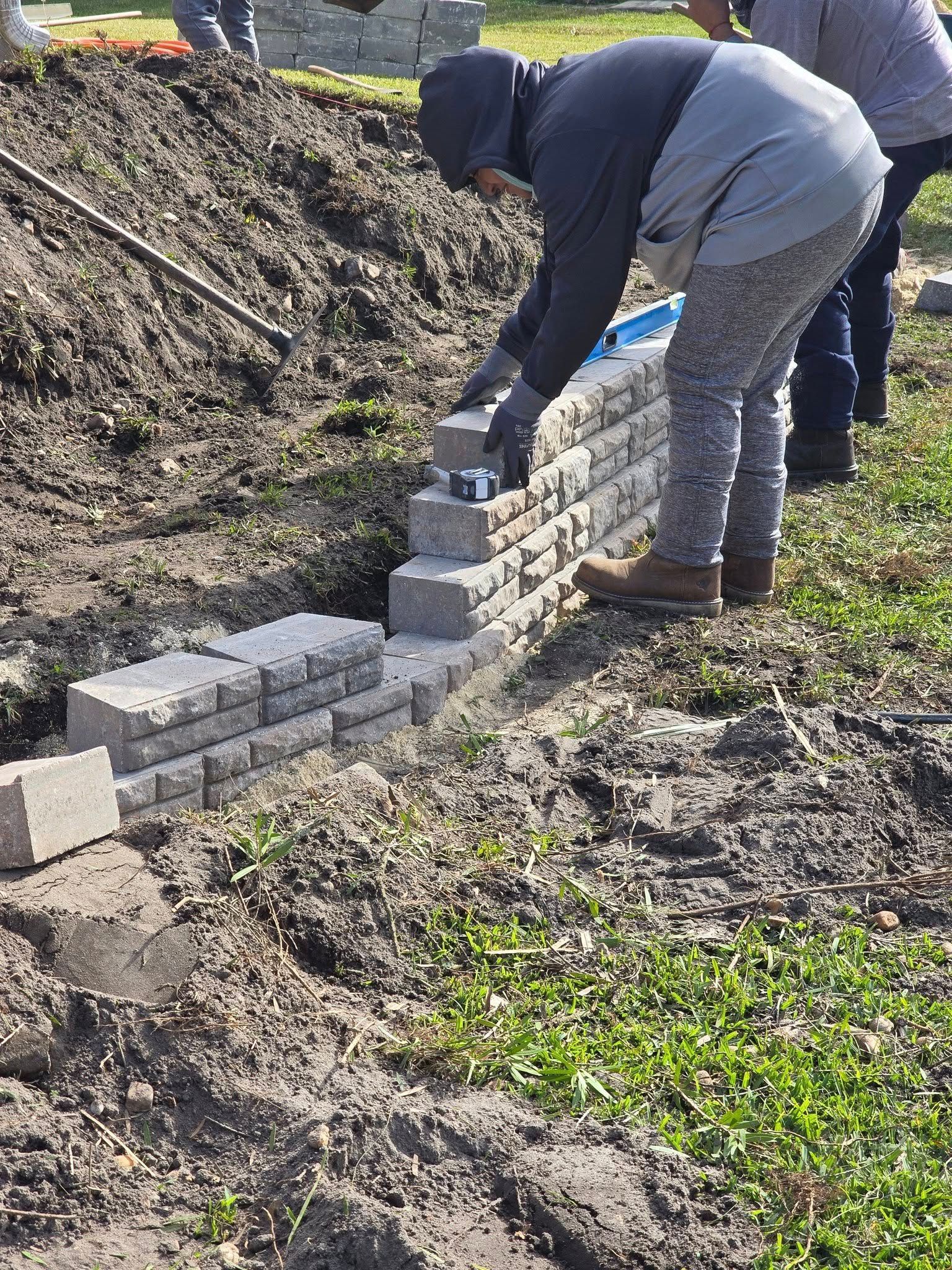 A person wearing a hoodie and patterned pants builds a stone retaining wall in a dirt-filled outdoor area.