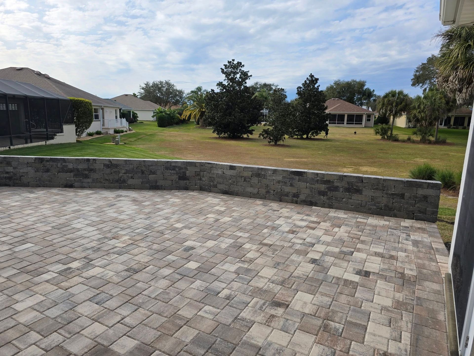 A stone paver patio features a low, gray stacked-stone retaining wall overlooking a grassy yard with trees and houses.