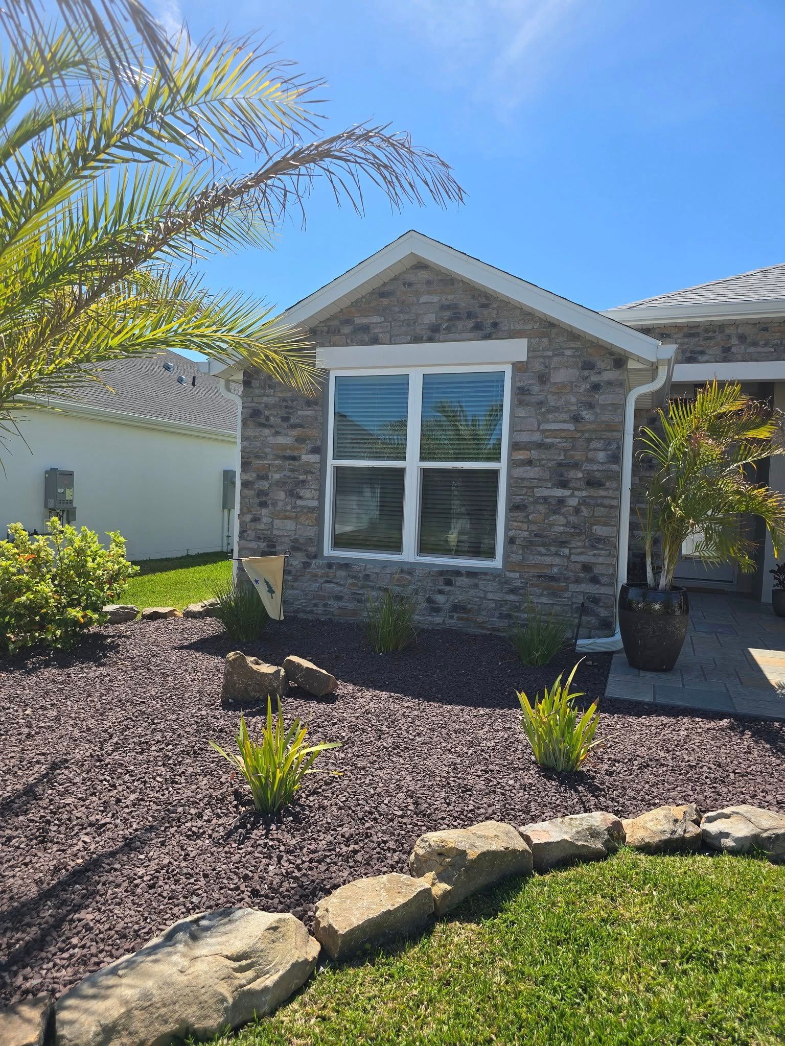 A stone-faced house exterior featuring a front window, dark landscaping rocks, and a rock-bordered lawn under a blue sky.