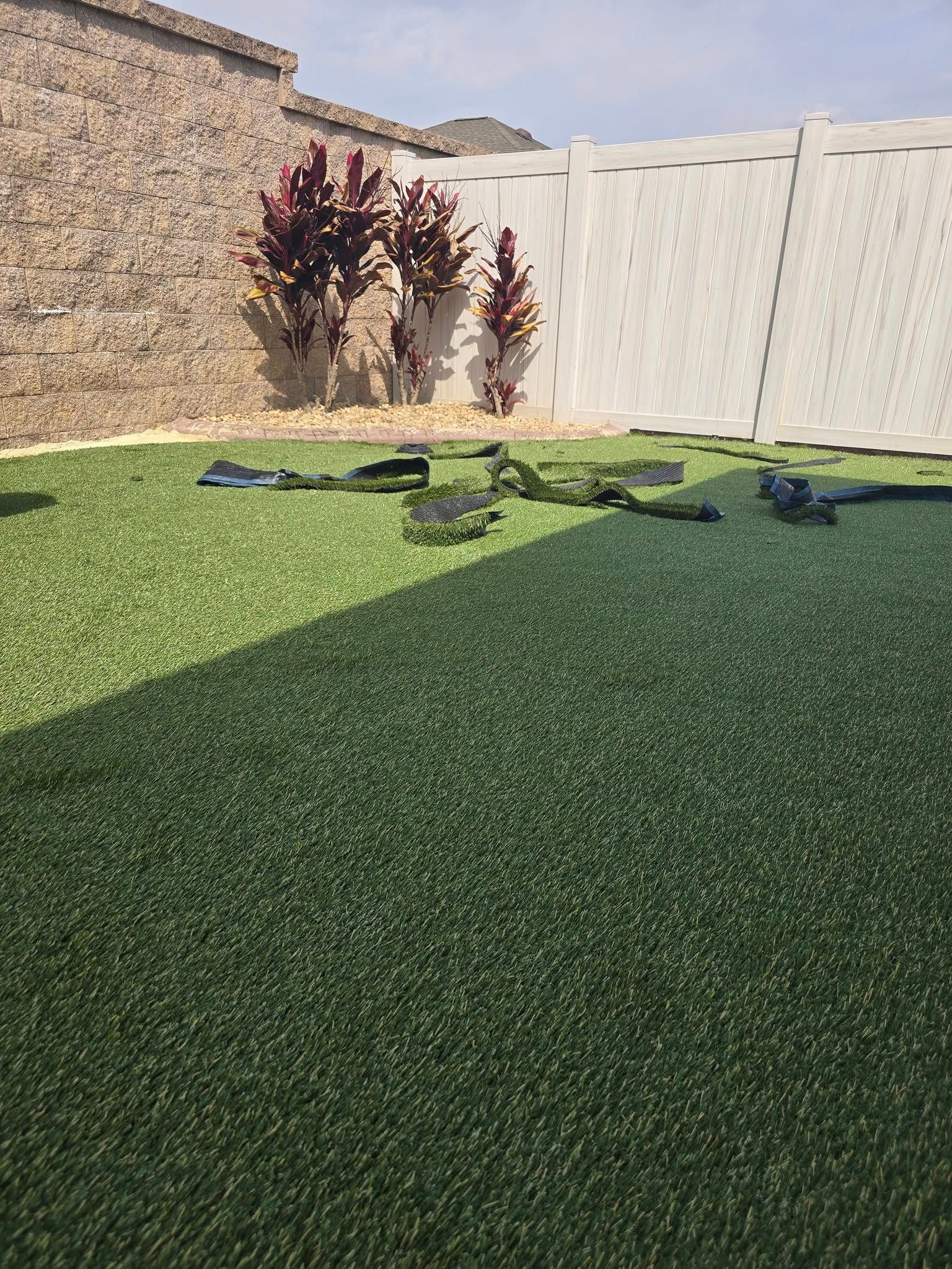 A backyard with artificial turf, a beige stone wall, a white vinyl fence, and three reddish-brown plants in the corner.