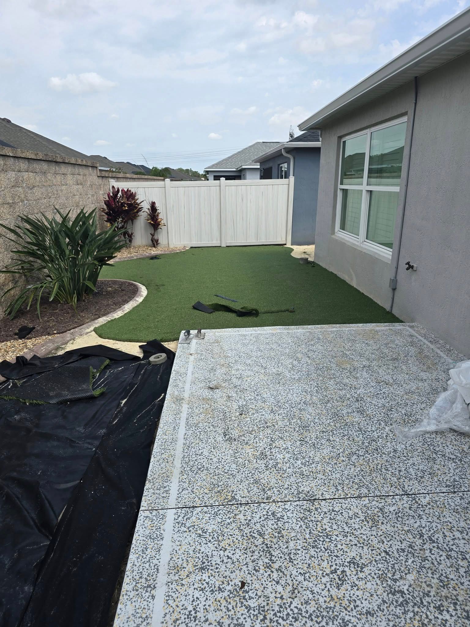 A backyard featuring a speckled patio, a section of green artificial turf, a white fence, and a gray house exterior.