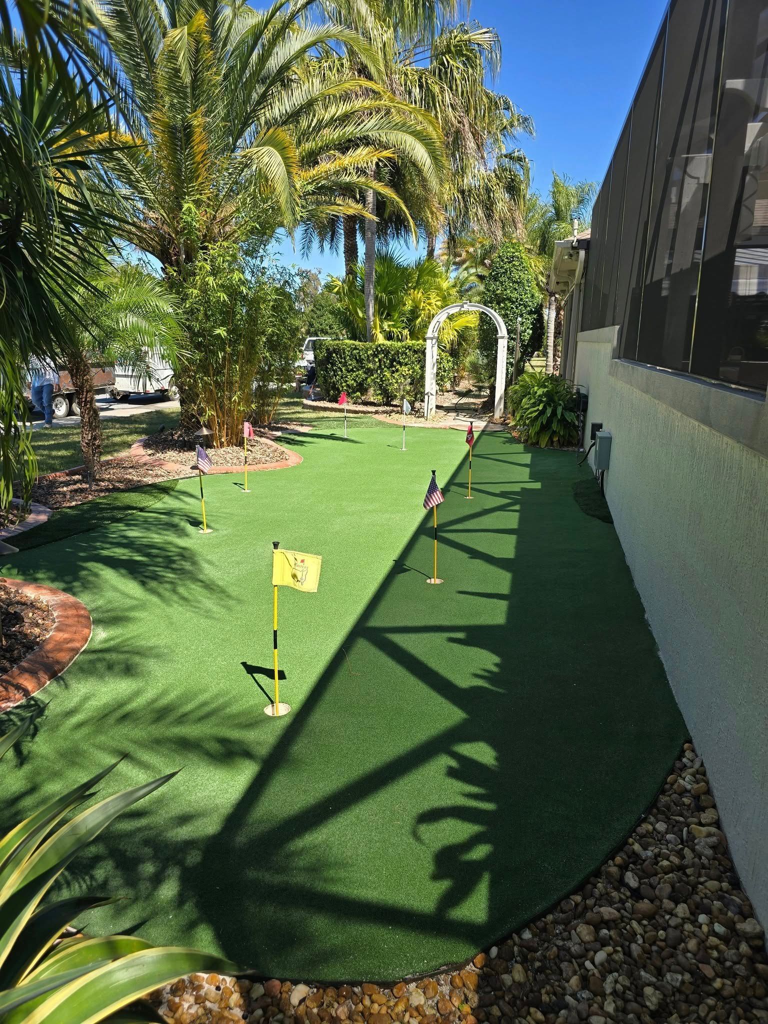 A small, backyard artificial grass putting green with three flagsticks, surrounded by palm trees and landscaping.