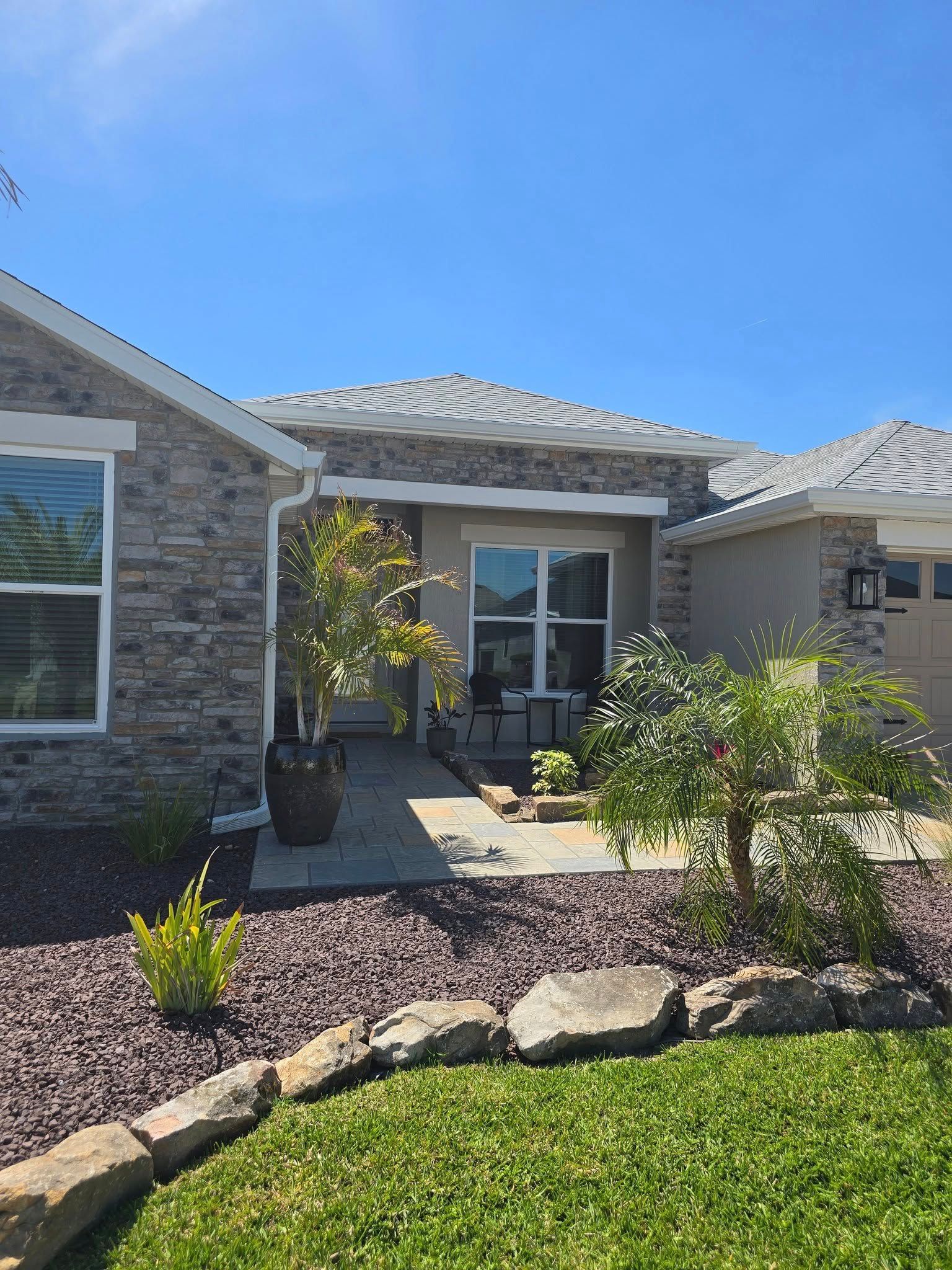 The front exterior of a stone-clad house under a clear blue sky, featuring a porch entry with small potted plants.