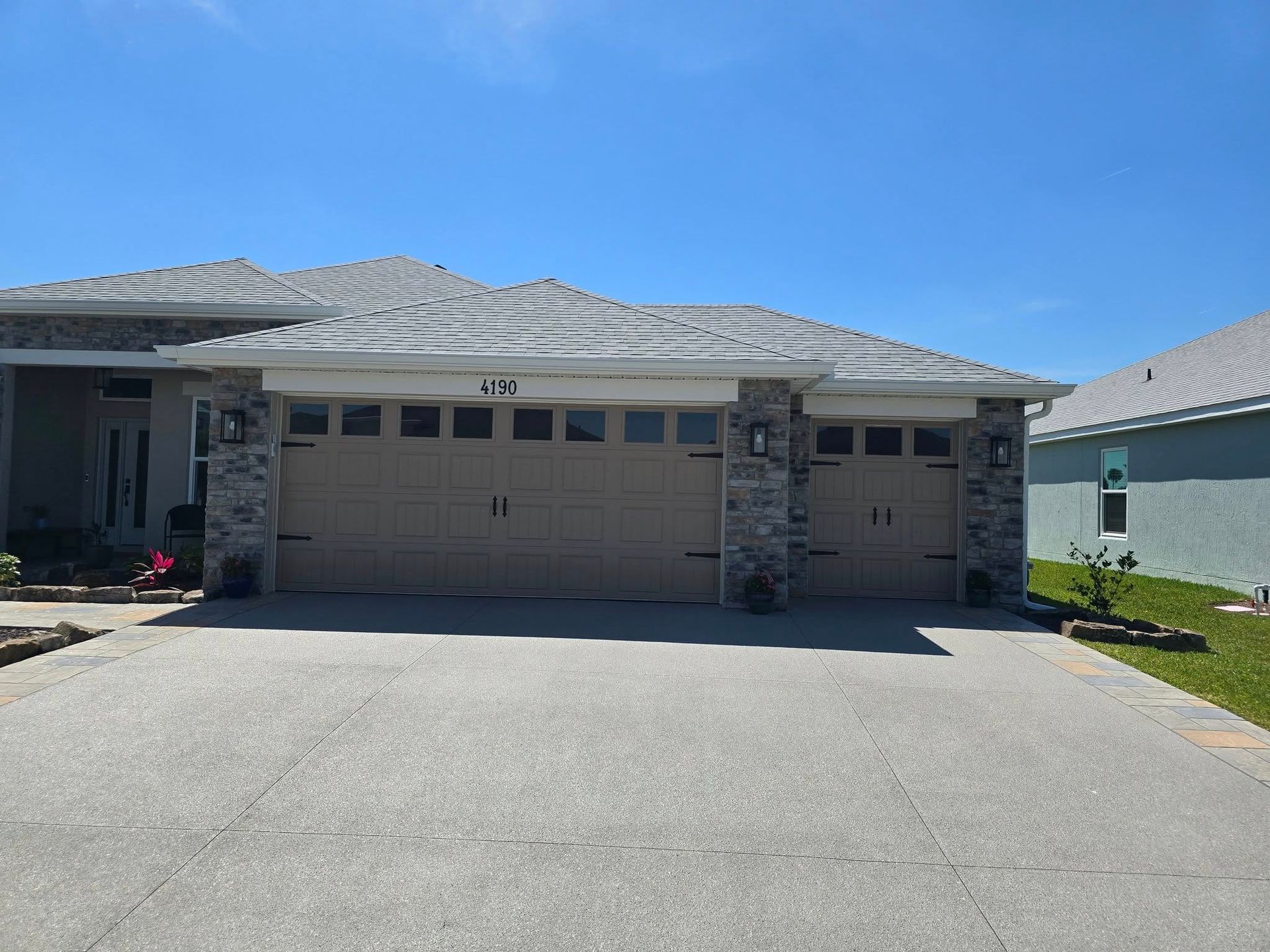 A beige, multi-car garage with stone siding and a light-colored roof, viewed from a residential concrete driveway.
