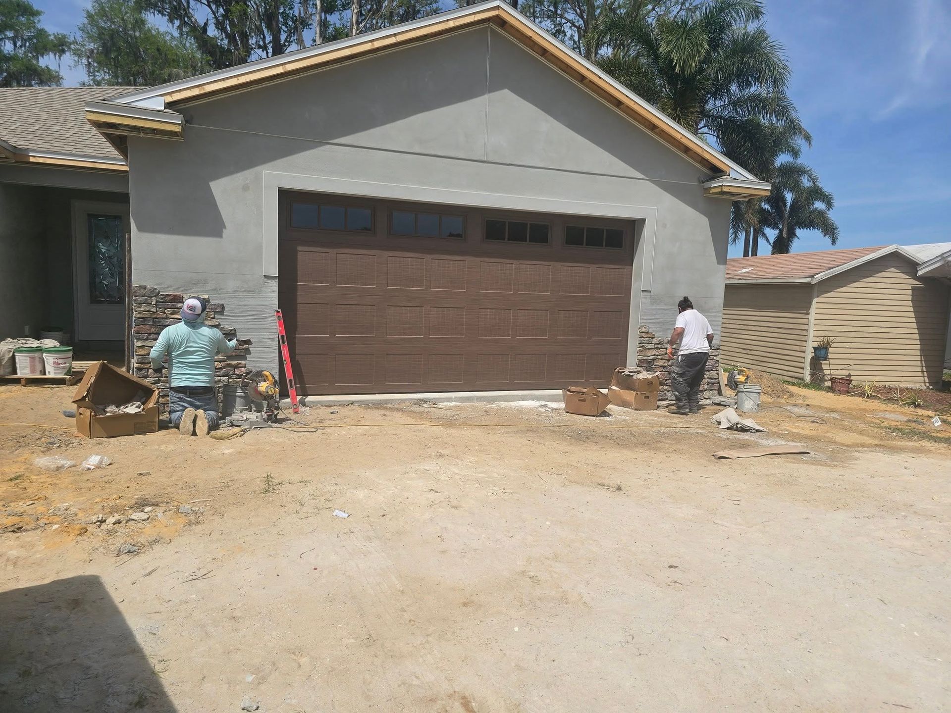 Two workers install stone veneer on the exterior wall of a house with a brown garage door.
