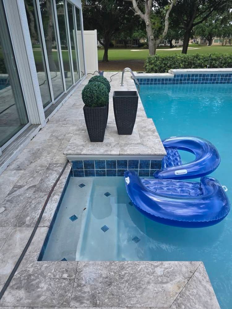 A blue inflatable pool float sits near the stone stairs of a swimming pool, next to two dark planters on a stone patio.