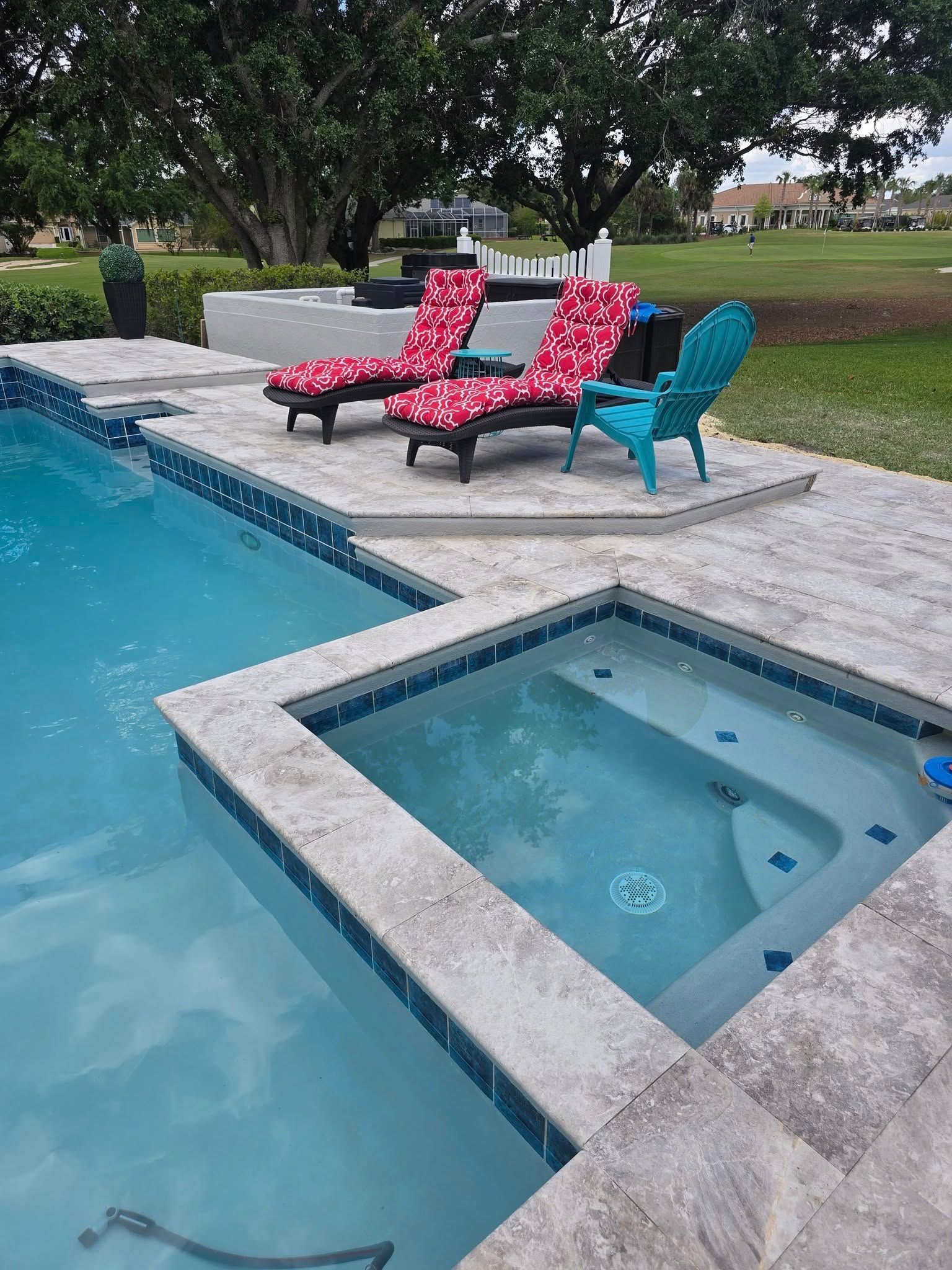 A backyard swimming pool with an attached spa, stone decking, two red floral lounge chairs, and a teal Adirondack chair.