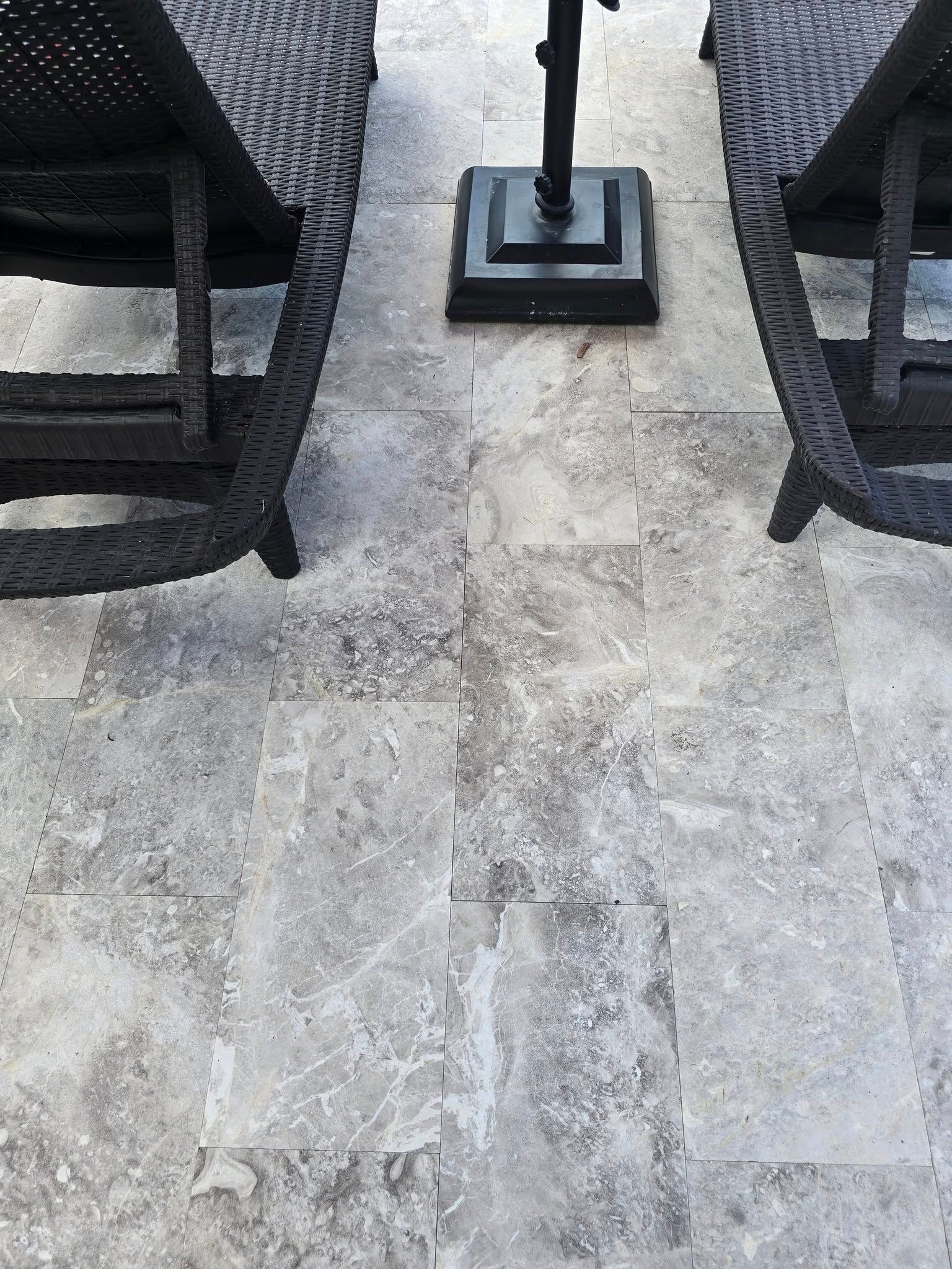 Two black outdoor lounge chairs flank a metal umbrella stand on a textured, grey stone patio.
