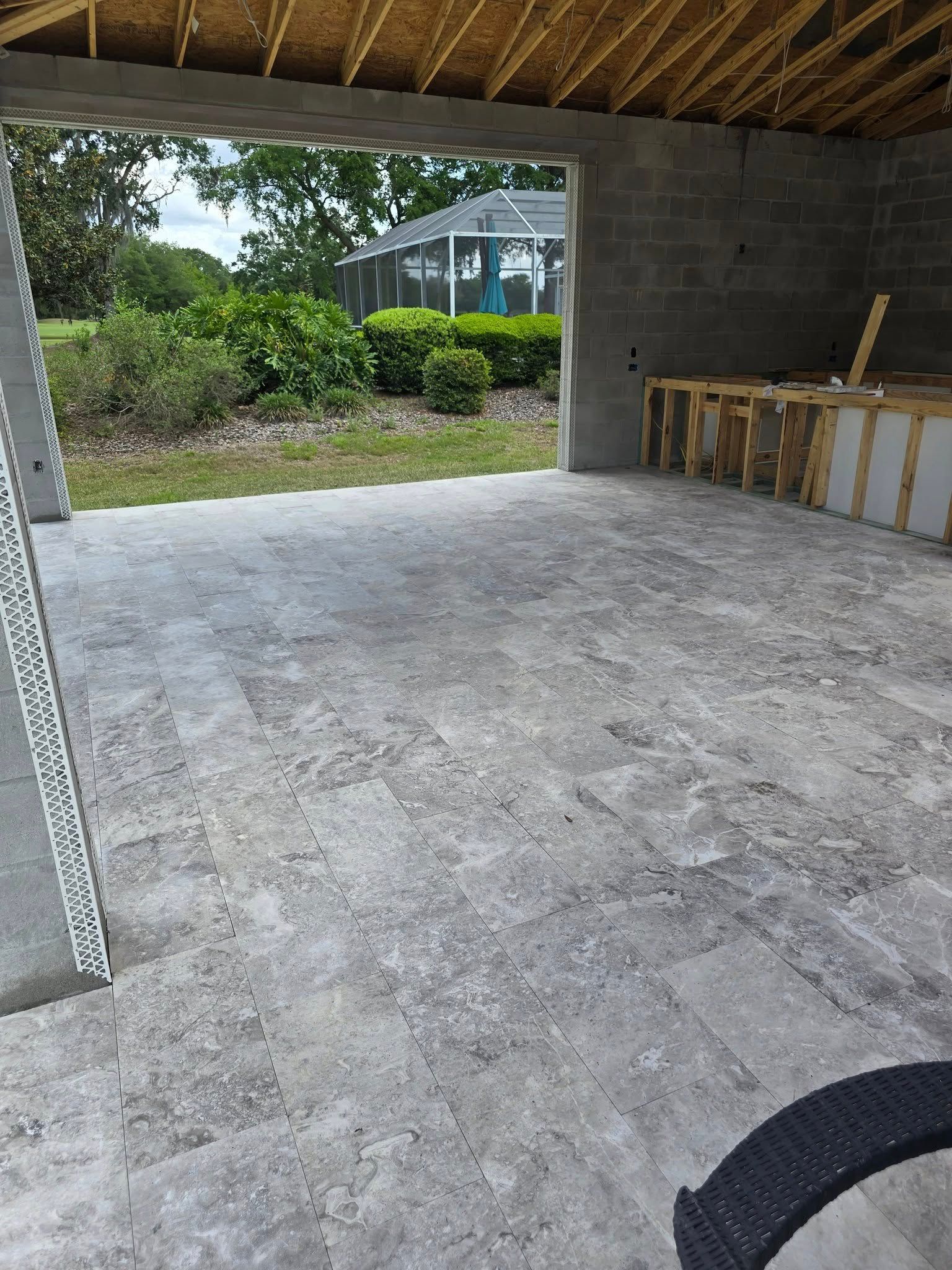 A partially constructed open-air room with gray stone tile flooring looking out toward a backyard and pool enclosure.