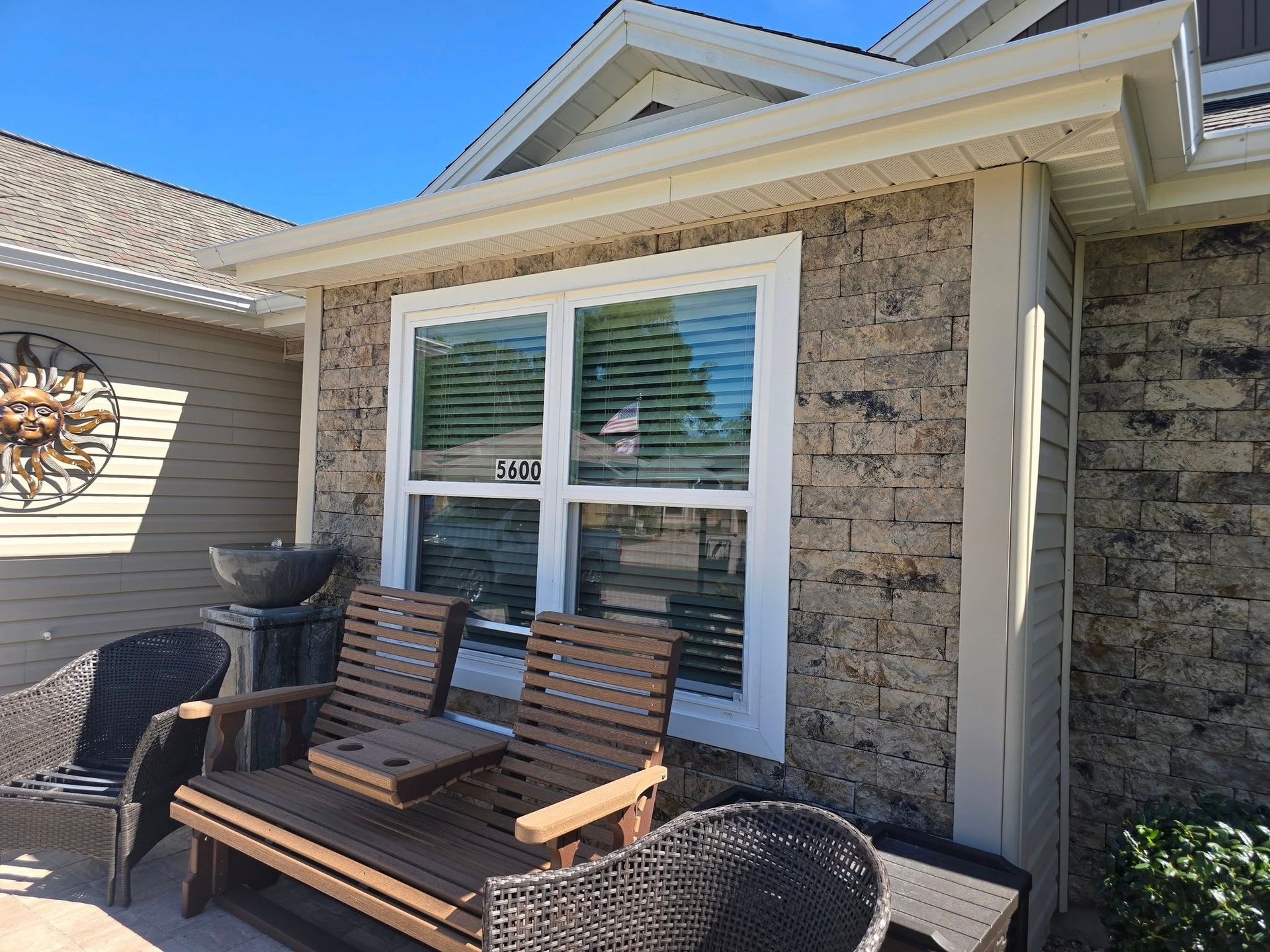 A stone-front house exterior with a double-pane window, patio furniture, and a decorative sun wall ornament.