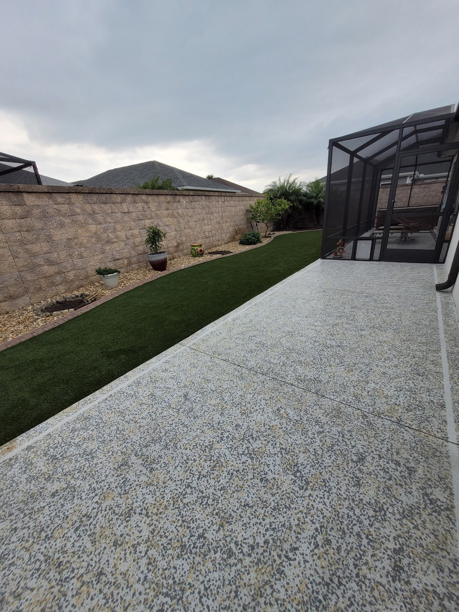 A patterned stone patio adjacent to a narrow strip of grass, bordered by a stone wall under a cloudy sky.