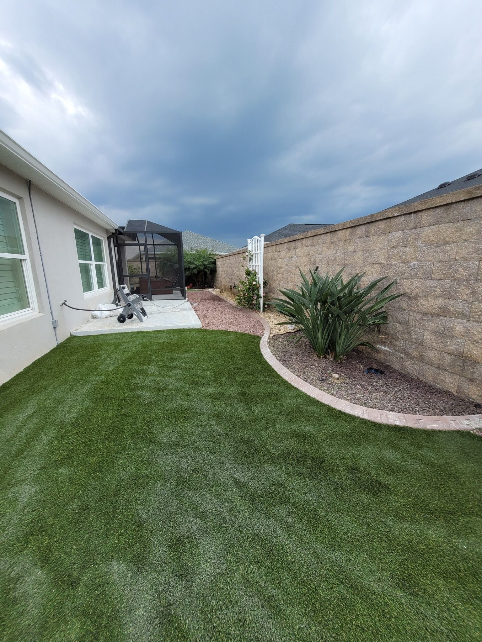 A side yard with a manicured green lawn, a landscaped garden bed with a bush, and a house wall under a cloudy sky.