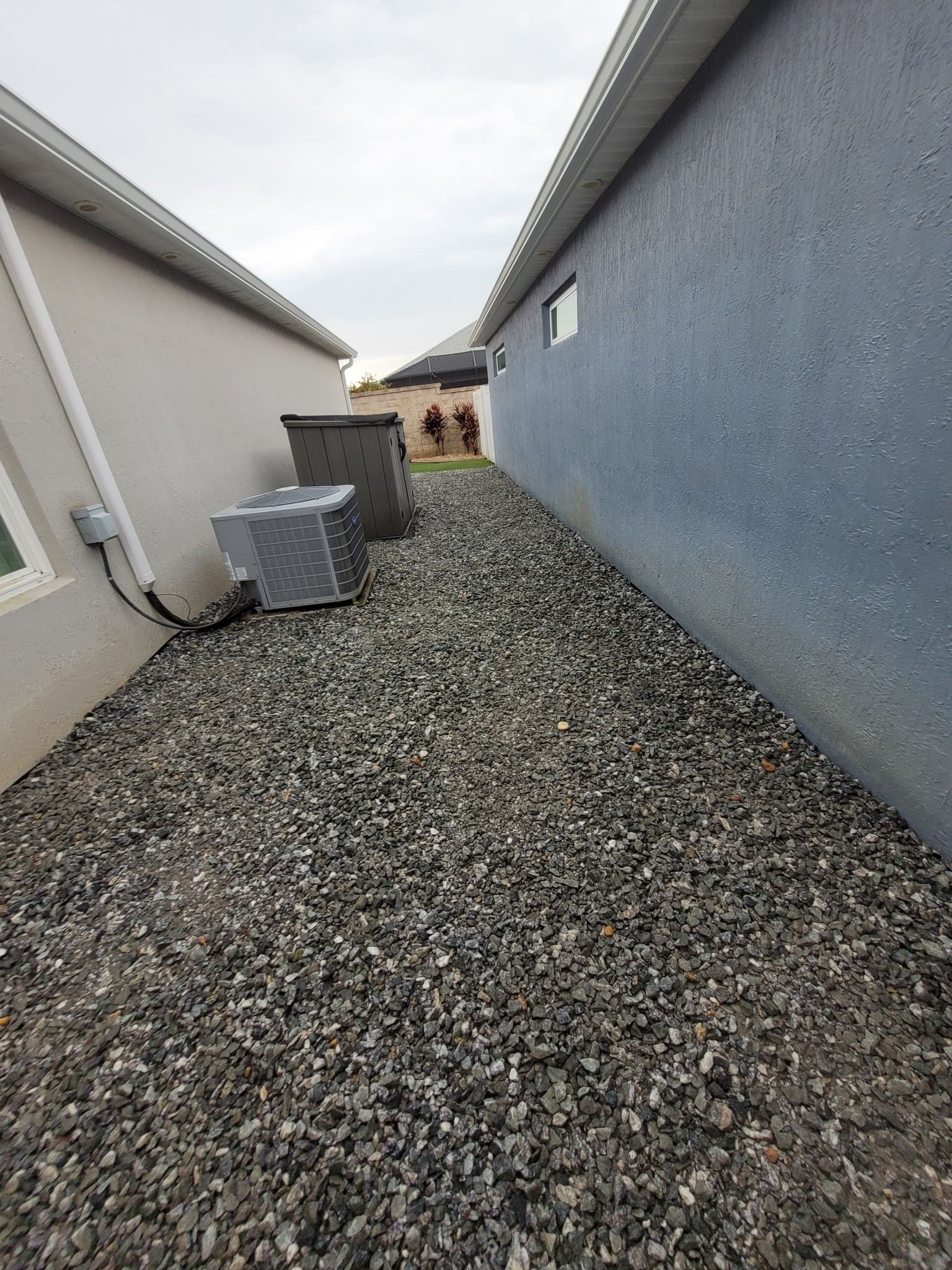A narrow gravel walkway between two house exterior walls, featuring an air conditioning unit and a trash bin.