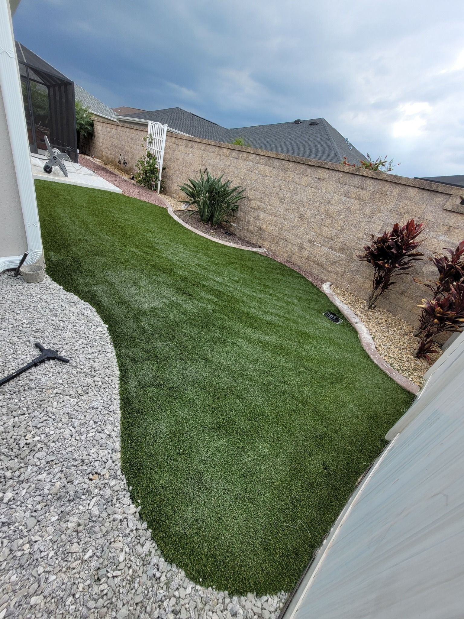 A backyard with a curved patch of artificial grass bordered by grey gravel, a stone wall, and some small shrubs.