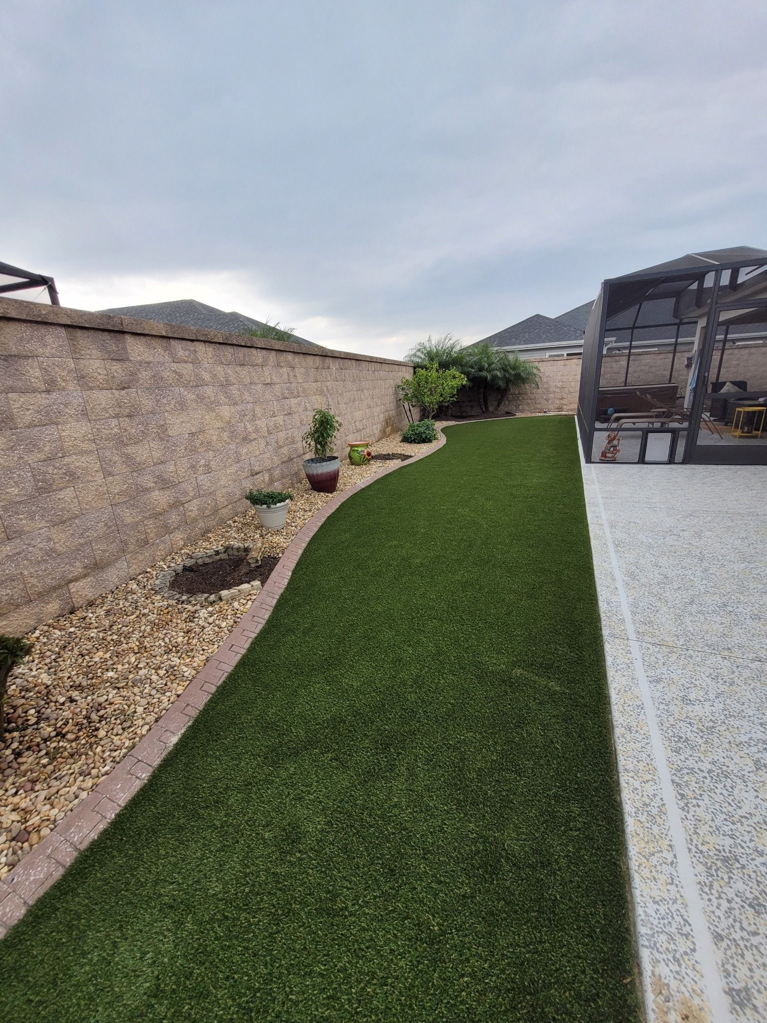 A backyard featuring a strip of green artificial turf, a rock garden border along a stone wall, and a paved patio area.