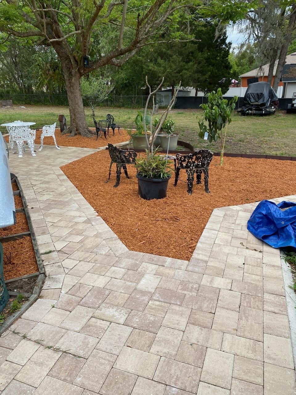 A paver patio walkway winds through a yard featuring orange wood mulch, decorative goat sculptures, and a large tree.