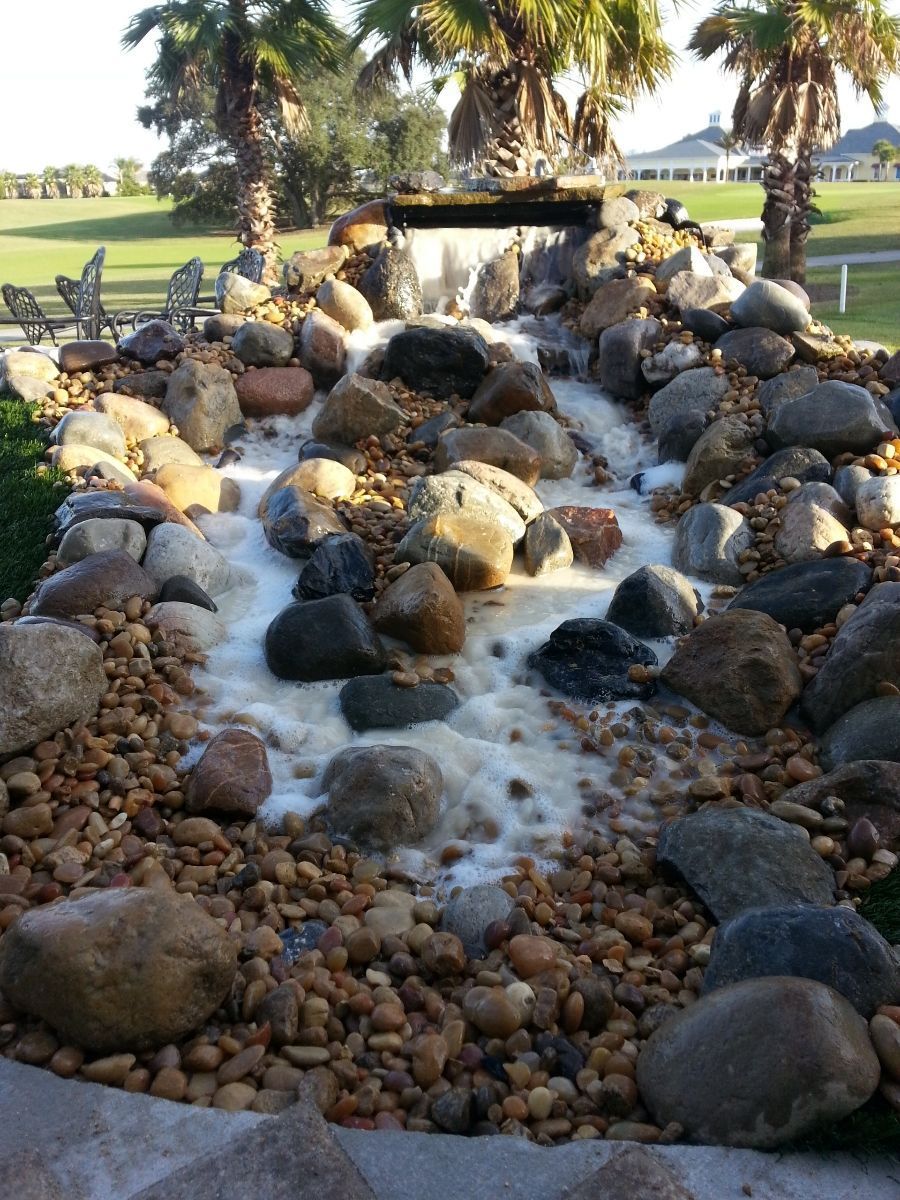 A tiered stone water feature flowing into a rocky stream in a palm-lined outdoor setting.