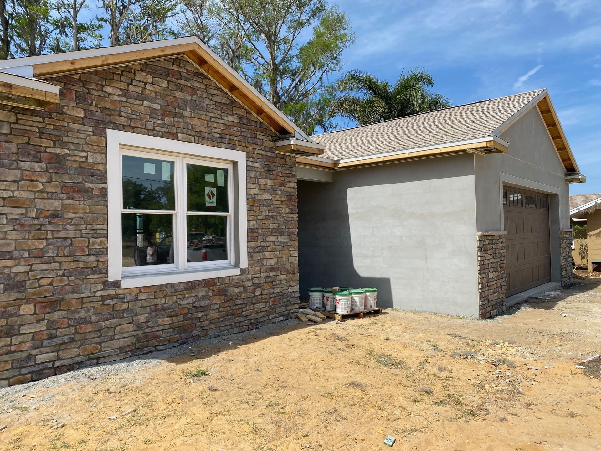 A partially constructed house with stone siding on the left, a grey stucco garage on the right, and a sandy dirt yard.