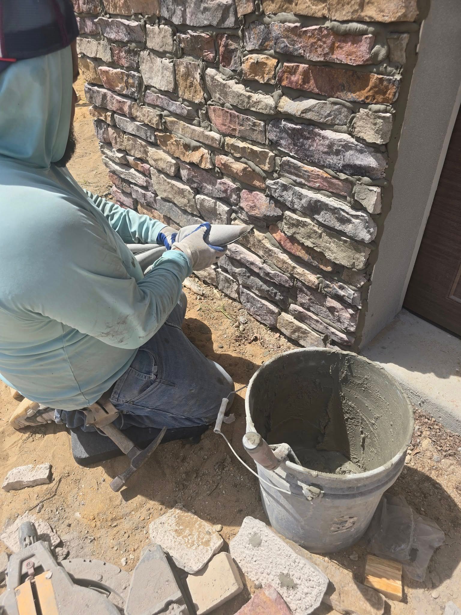 A person uses a grout bag to fill the spaces between stones on a decorative exterior masonry wall.