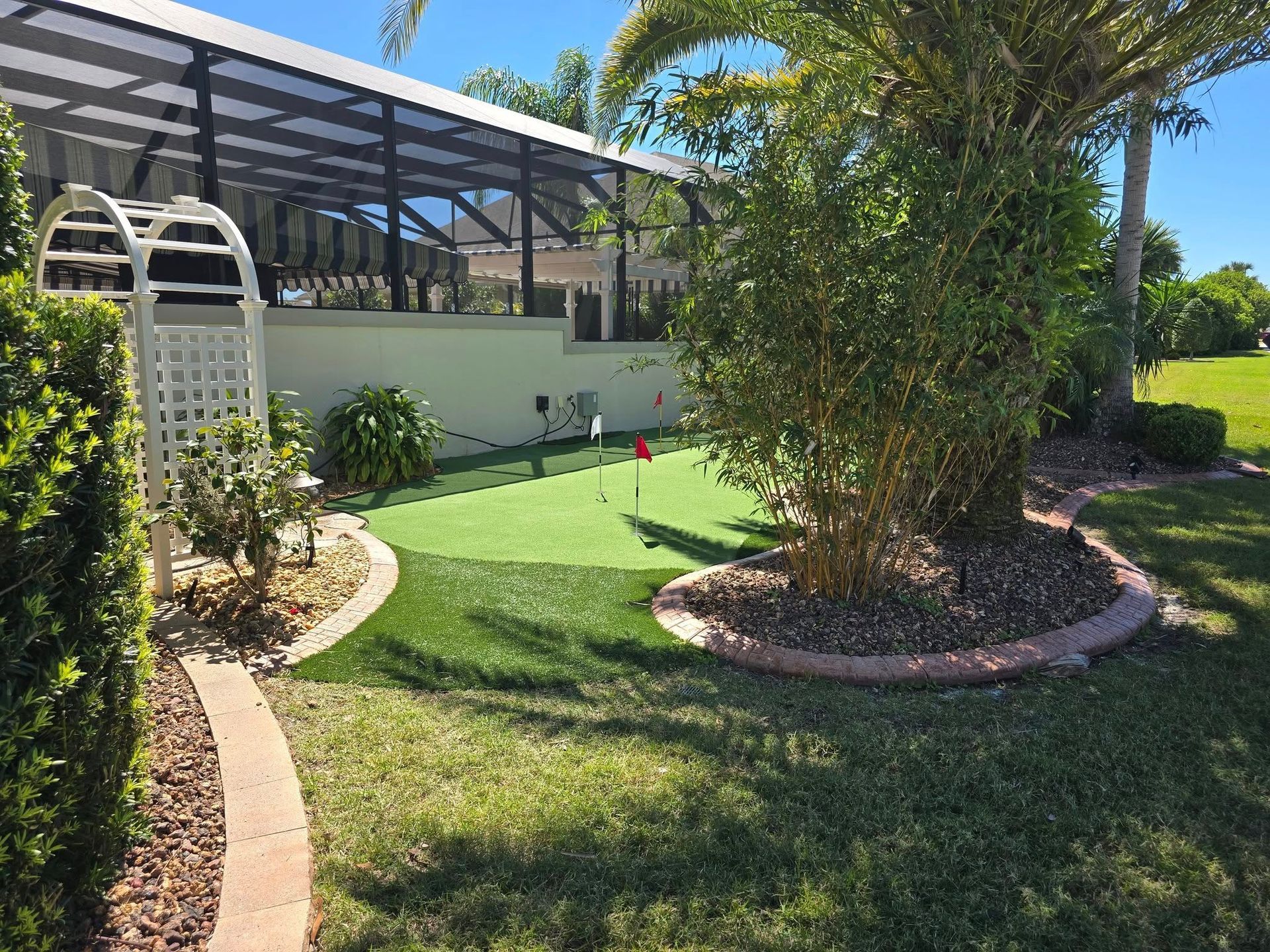 A small backyard putting green with a white trellis archway, bordered by mulch beds and a palm tree under a sunny sky.