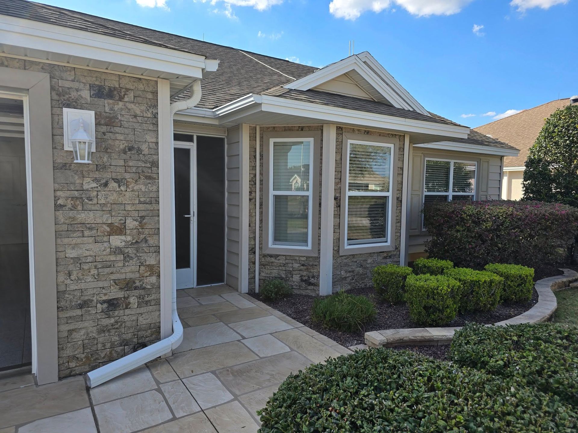 A single-story home exterior with beige stone siding, a tiled walkway, a front entryway, and pruned green hedges.