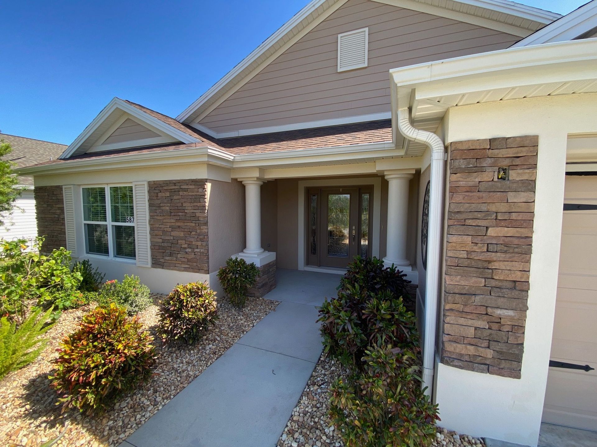 A tan, single-story house with a stone-accented exterior, white columns at the entrance, and a landscaped front yard.