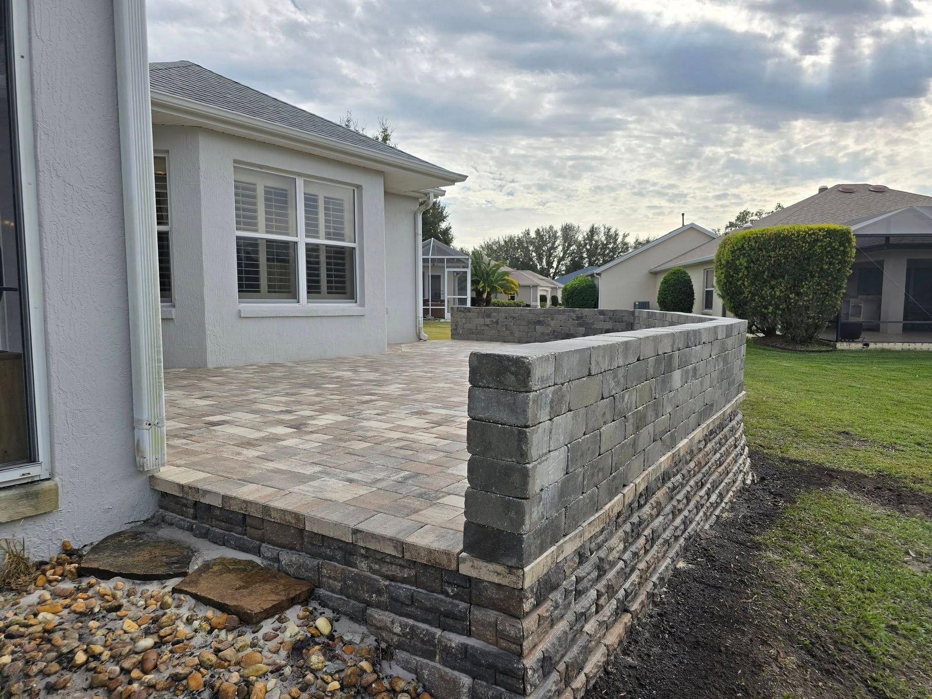 A stone paver patio with a low, decorative retaining wall in a grassy residential backyard.